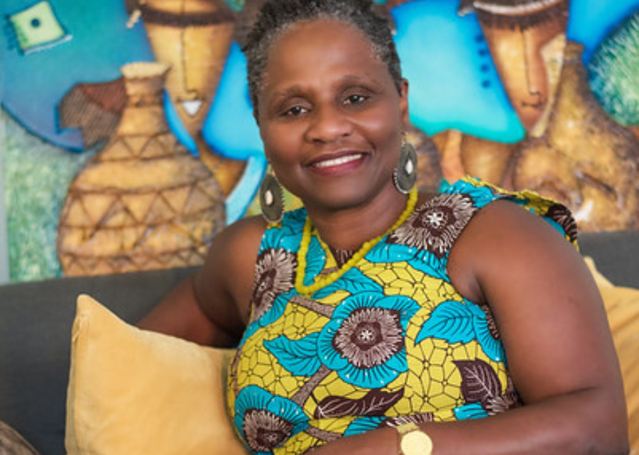 Licensed Mental Health Therapist Jacqueline Samuda is a black woman with short natural black and gray hair, dark brown skin, brown eyes, red lipstick. She is wearing a Jamaican dress with turquoise, mustard, brown and white design. She is sitting on a brown sofa with mustard color pillows.